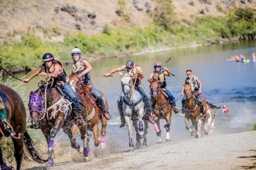 A group of riders on horseback gallops along a riverbank, splashing water as they race in a lively outdoor event.