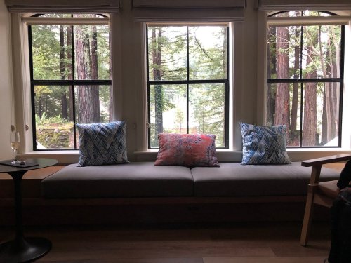 Cozy window seat with colorful pillows, overlooking a forested view, and a glass of champagne on a small table.