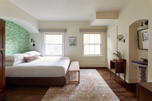 Bright, modern bedroom with a large bed, green patterned wall, wooden furniture, and natural light from two windows.