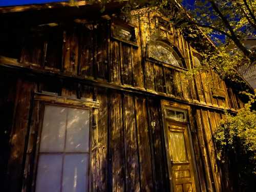 A weathered wooden house at night, with shadows cast by nearby trees and a partially visible door and window.