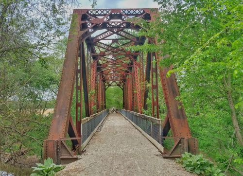 A rusty metal bridge spans a tree-lined path, with greenery surrounding the structure and a bicycle parked on the bridge.