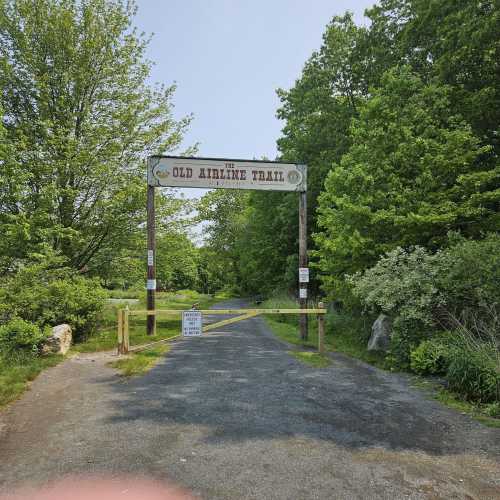 A wooden sign marks the entrance to the Old Airline Trail, surrounded by greenery and a clear blue sky.