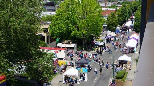 A bustling street fair with vendors, tents, and crowds under sunny skies, surrounded by trees and buildings.