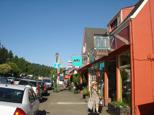 A sunny street scene with shops, colorful signs, and a person walking along the sidewalk in a quaint town.