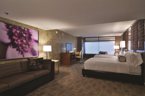 A modern hotel room featuring two beds, a sofa, a desk, and large windows with decorative wall art.