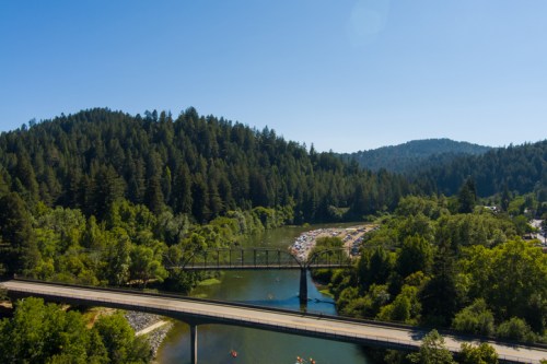 A scenic view of a river surrounded by lush green trees, featuring a bridge and a parking area in the distance.