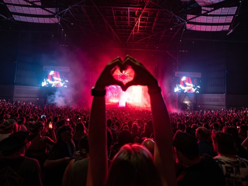 A crowd at a concert with a person making a heart shape with their hands against a vibrant, colorful backdrop.