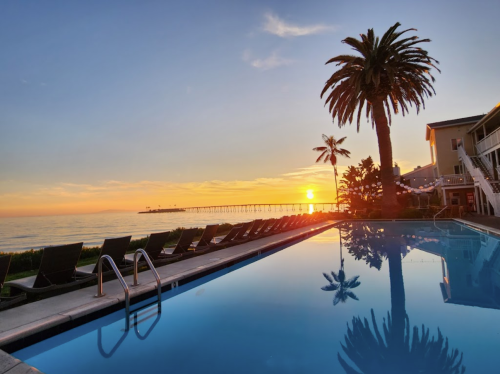 Sunset over a calm ocean, reflecting in a pool with palm trees and a pier in the background.