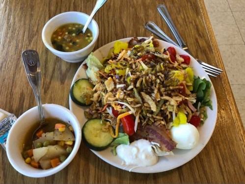 A large salad topped with crispy onions, tomatoes, and eggs, served with a side of soup and cucumbers on a wooden table.
