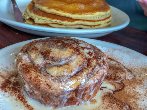 A cinnamon roll drizzled with icing, with a stack of pancakes in the background on a plate.