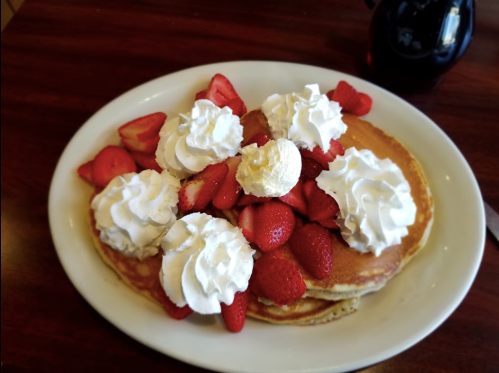 A plate of pancakes topped with whipped cream and fresh strawberries, served with a small pitcher of syrup.
