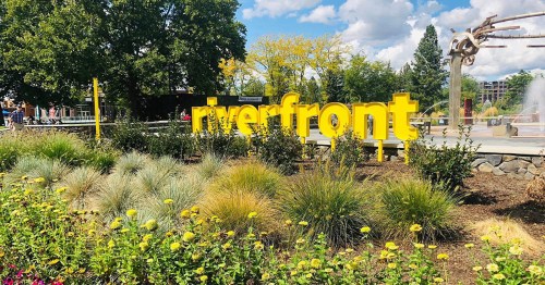 A vibrant sign reading "riverfront" surrounded by colorful flowers and greenery under a blue sky with clouds.