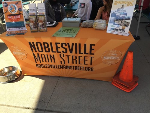 A table with a bright orange tablecloth displaying "Noblesville Main Street" and promotional materials at an outdoor event.