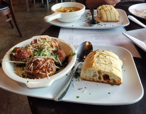 A plate with three meatballs topped with cheese, a side of bread, and a bowl of soup in the background.