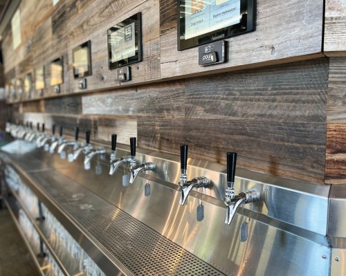 A row of beer taps on a stainless steel counter against a wooden wall, with digital screens above each tap.