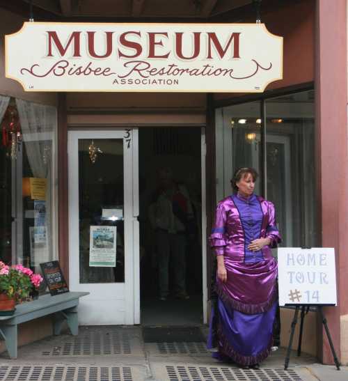 A woman in a purple Victorian dress stands outside a museum entrance, with a sign for a home tour nearby.