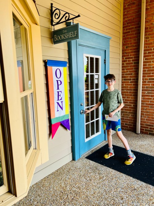 A child stands at the entrance of a bookstore, holding a book, with an "OPEN" sign displayed.