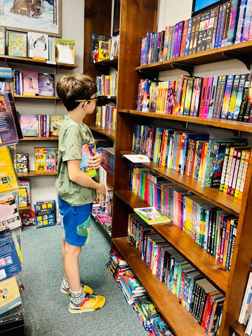 A child in a green shirt and shorts browses colorful books on a shelf in a cozy bookstore.