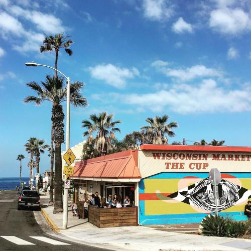 Colorful beachside market with palm trees, people sitting outside, and ocean in the background under a blue sky.