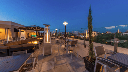 Rooftop terrace with seating, heaters, and a view of a city skyline at dusk.