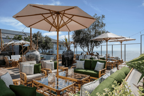 A stylish outdoor lounge with green seating, umbrellas, and ocean views, surrounded by plants and decorative elements.