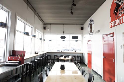 Bright, modern restaurant interior with a long wooden table, black stools, and large windows. Red doors and decor visible.