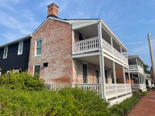 Historic brick house with white balconies, surrounded by greenery and a blue sky in the background.