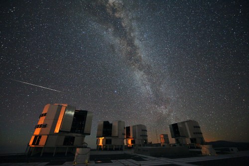 A starry night sky with the Milky Way above observatory buildings, illuminated by soft orange lights.