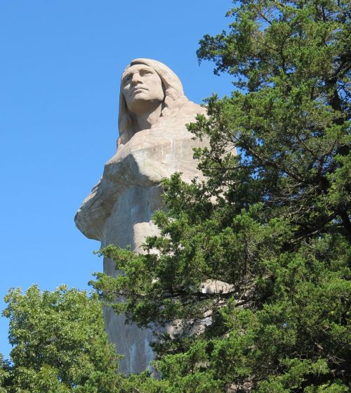 A tall stone statue of a Native American figure surrounded by trees against a clear blue sky.