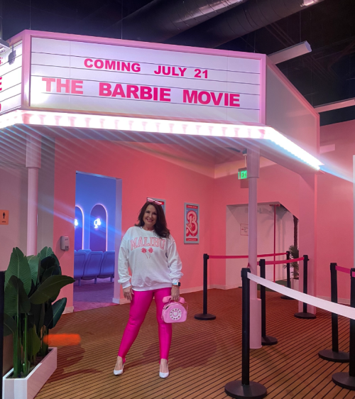A person in a pink-themed space poses in front of a movie marquee for "The Barbie Movie," wearing a white sweatshirt and pink leggings.