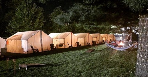 A row of illuminated tents at night, with a hammock and a fire pit in a grassy area surrounded by trees.
