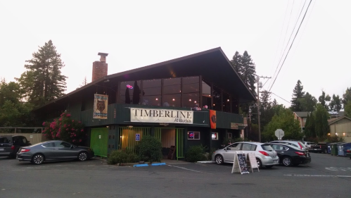 A green building with large windows, "Timberline" sign, surrounded by trees and parked cars in a quiet area.