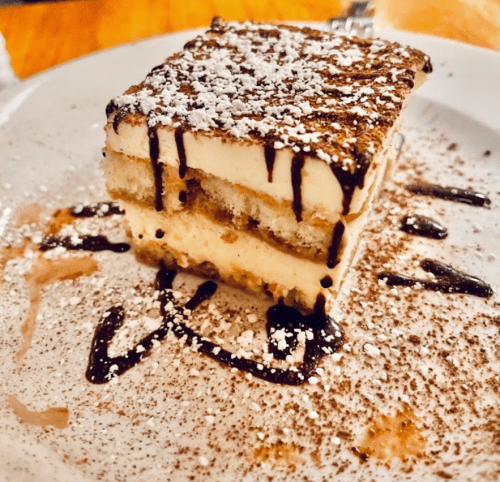 A slice of tiramisu dusted with powdered sugar and drizzled with chocolate sauce on a white plate.