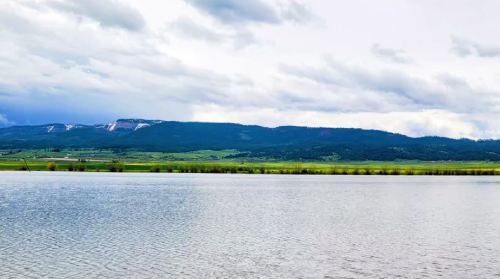 A serene lake reflects a cloudy sky, with lush green hills and distant mountains in the background.