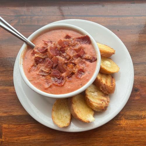 A bowl of creamy tomato soup topped with crispy bacon, served with golden-brown toasted bread on a plate.