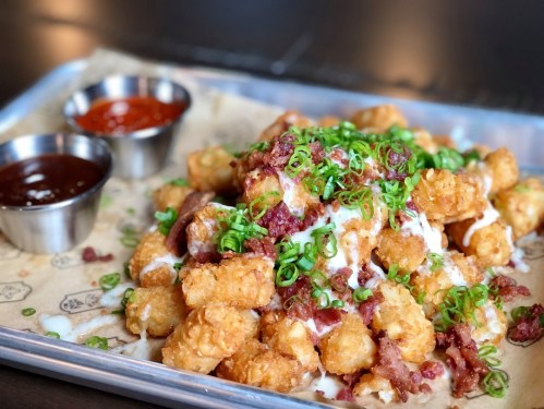 A tray of loaded tater tots topped with green onions, drizzled with sauce, and served with two dipping sauces.