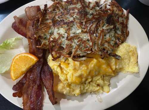 A plate of breakfast featuring crispy hash browns, scrambled eggs with cheese, bacon, and a slice of orange.