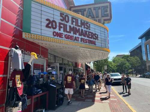 A bustling street scene in front of a theater marquee advertising a film festival with crowds and merchandise for sale.