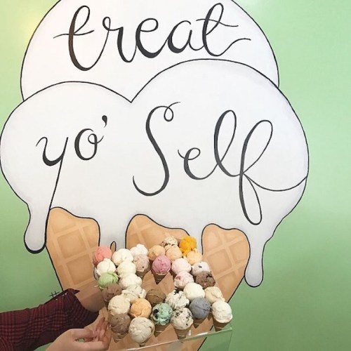 A hand holds a large ice cream cone filled with various colorful scoops, against a green wall with "treat yo' self" written above.