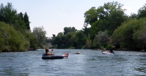 Two people relax on inner tubes in a calm river, surrounded by trees and greenery under a clear blue sky.