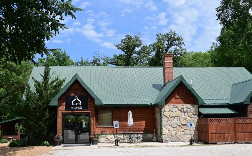 A rustic wooden building with a green metal roof, featuring a sign and outdoor seating under a tree.