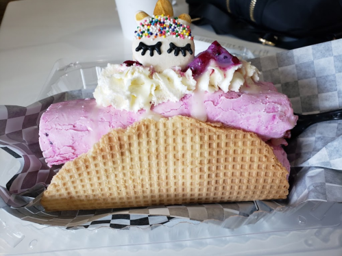 A colorful ice cream roll in a waffle cone, topped with whipped cream, sprinkles, and a cute unicorn decoration.