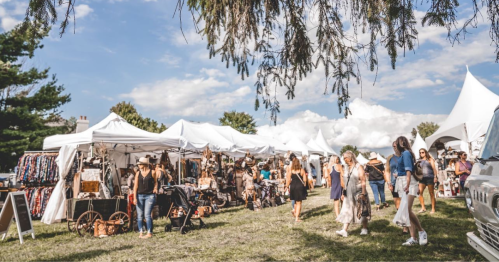 A bustling outdoor market with white tents, vendors, and shoppers enjoying a sunny day.
