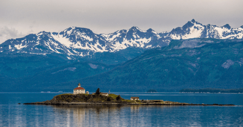 A serene island with a lighthouse surrounded by calm waters and snow-capped mountains in the background.