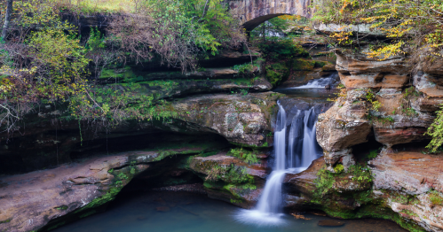 A serene waterfall cascades over rocky cliffs, surrounded by lush greenery and a stone bridge above.