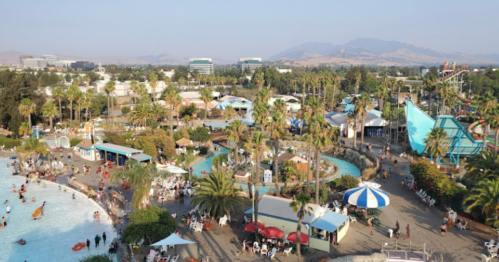 Aerial view of a vibrant water park with palm trees, pools, and attractions, set against a backdrop of mountains.