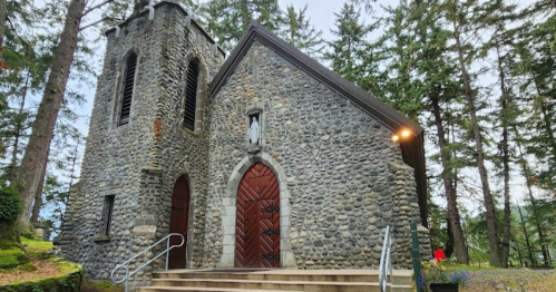 A stone church with wooden doors, surrounded by tall trees and greenery.