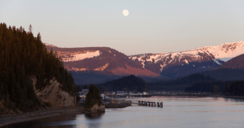 A serene landscape featuring a river, mountains with snow, and a full moon in the sky at dusk.