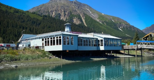 A waterfront building with large windows, surrounded by mountains and clear blue water on a sunny day.