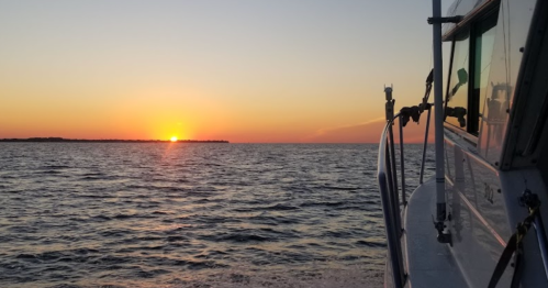 A boat is anchored near the shore as the sun sets over the calm ocean, casting warm colors in the sky.